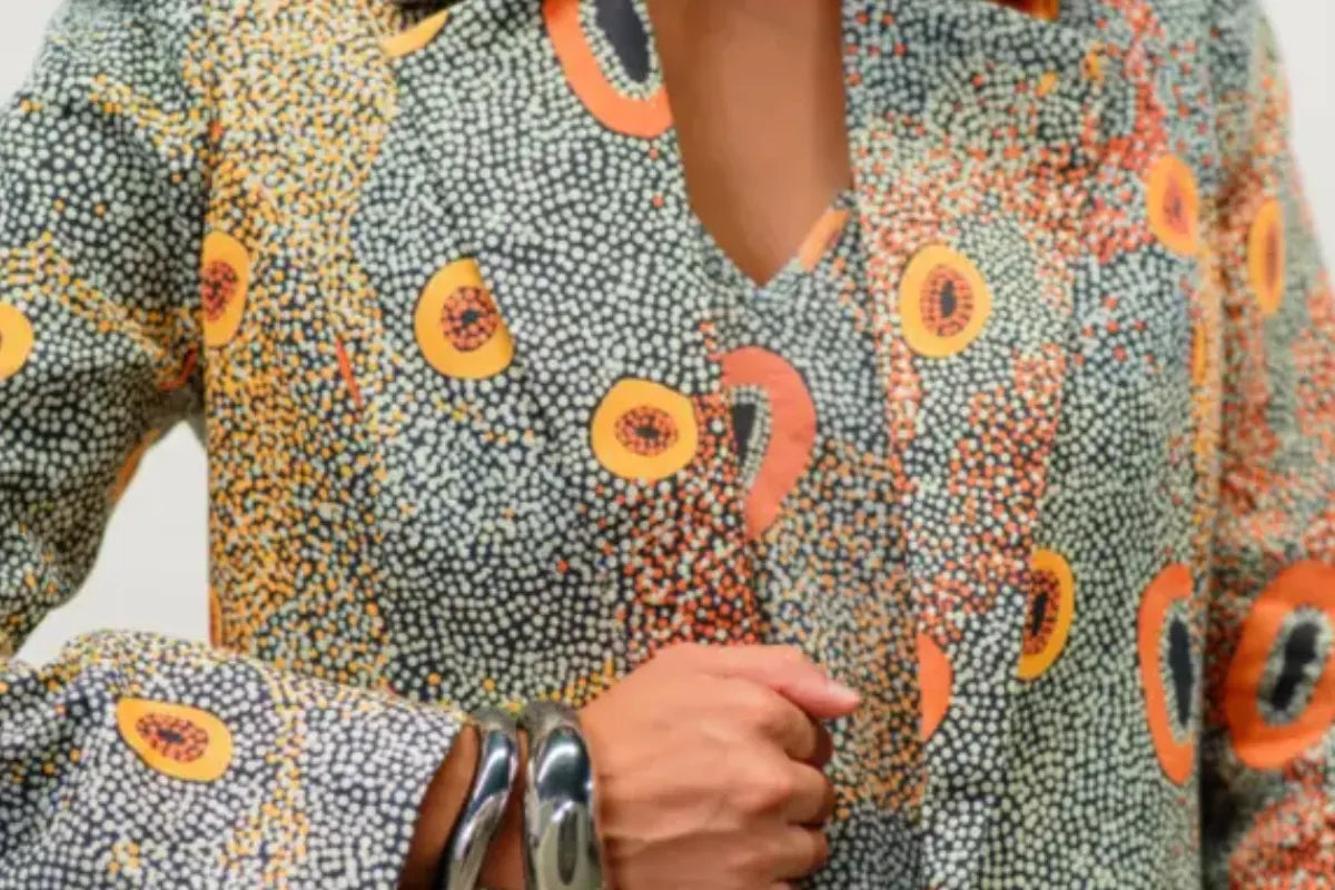 Close-up of a person wearing a Kirrikin long-sleeved V-neck top in the "Dry Country" print, featuring intricate Indigenous dot artwork and orange circular motifs, styled with thick silver bangles