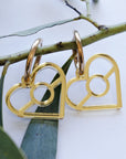 Aboriginal Gold Flag Heart Dangle Earrings hanging on a botanical branch, featuring metallic gold-toned heart cutout charms with a central ring design on gold hoop ear wires, designed by Buffie Punch.