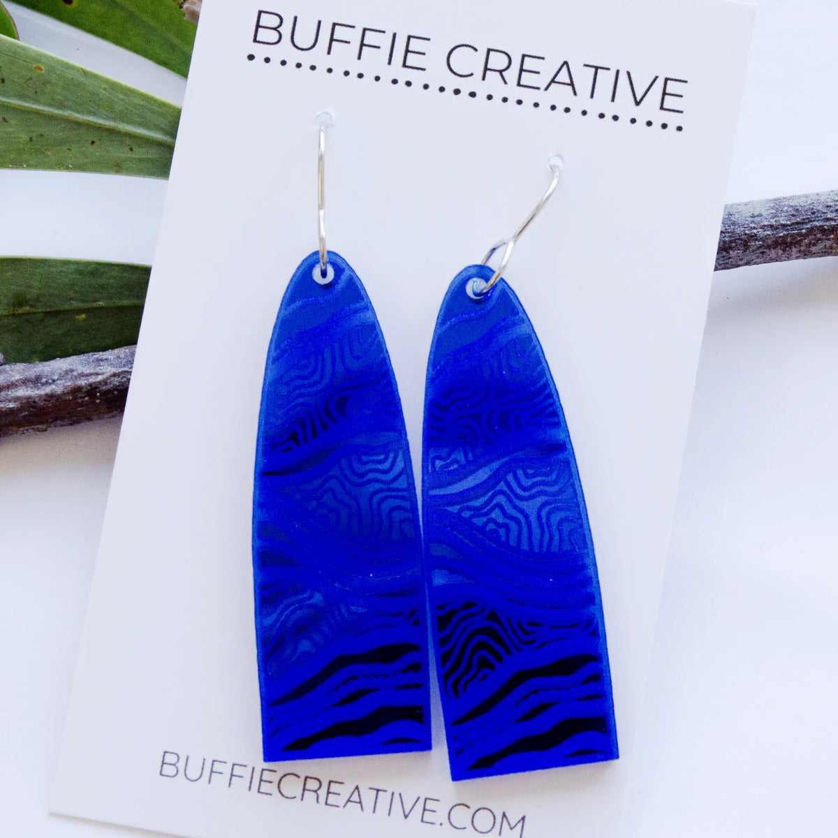 Bidi Blue Earrings on Buffie Creative card packaging, featuring elongated arch drops in vibrant electric blue with textured linework and pathway patterns on silver ear wires, designed by Buffie Punch.