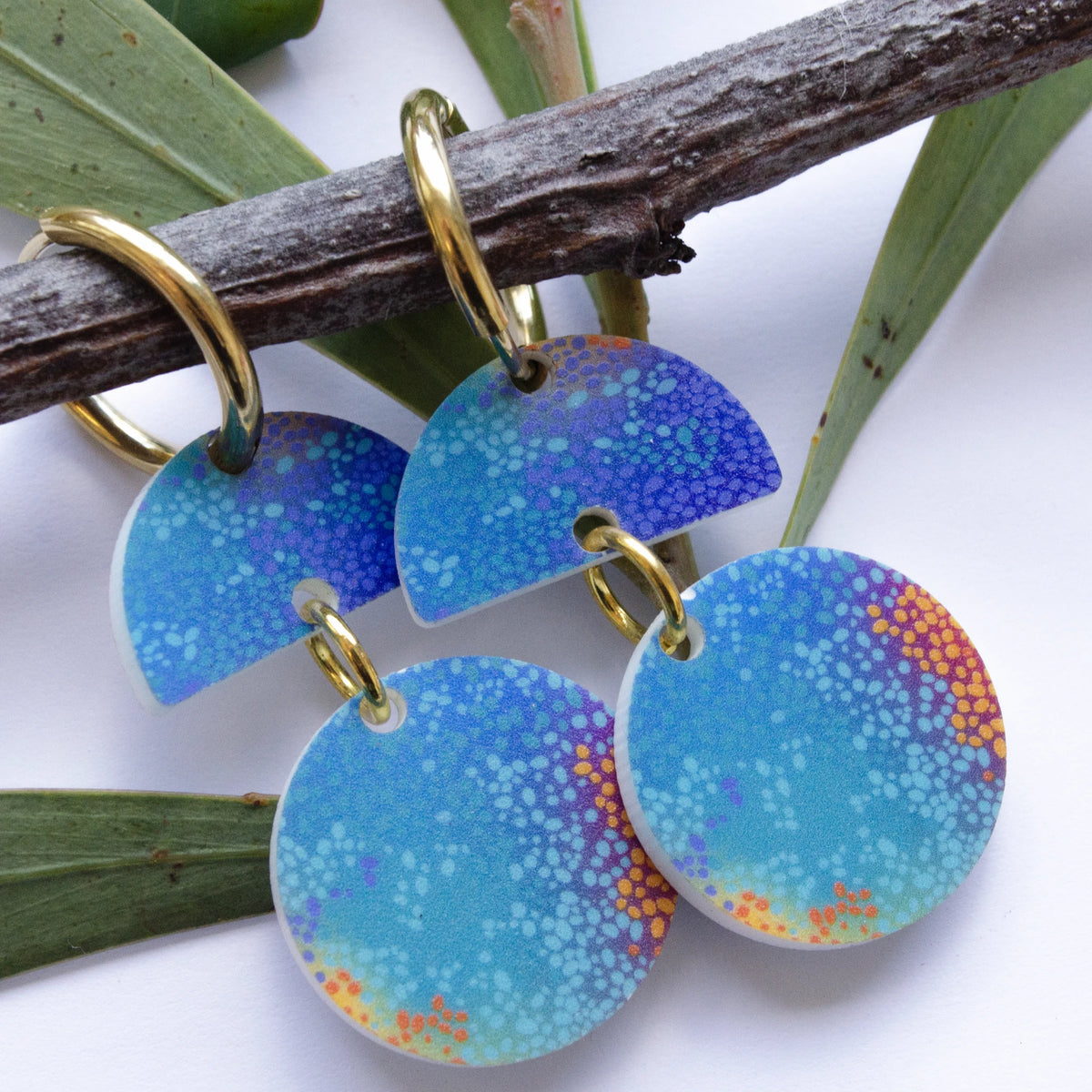 Blue Country Double Drop Earrings featuring articulated half-circle and full-circle charms with blue, turquoise, purple, and ochre Indigenous dot artwork hanging from gold-toned hoops, designed by Buffie Punch.