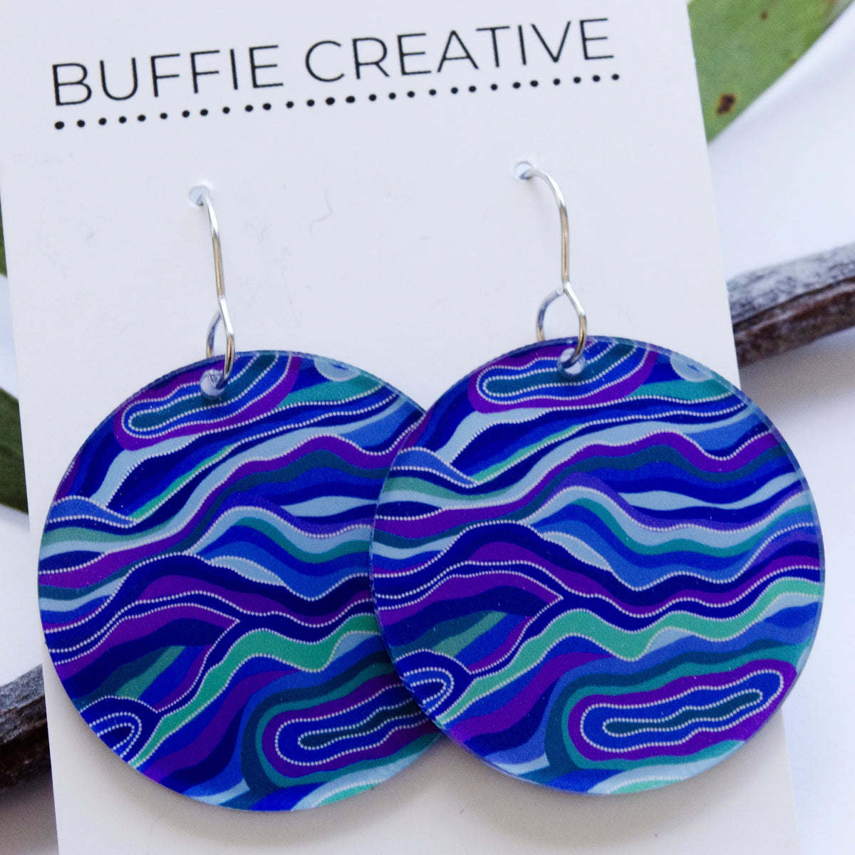 Blue &amp; Purple Circular Statement Earrings on Buffie Creative card packaging, featuring large round drop discs with flowing wavy line patterns in blue, purple, turquoise, and green Indigenous artwork on silver-toned ear wires, designed by Buffie Punch.