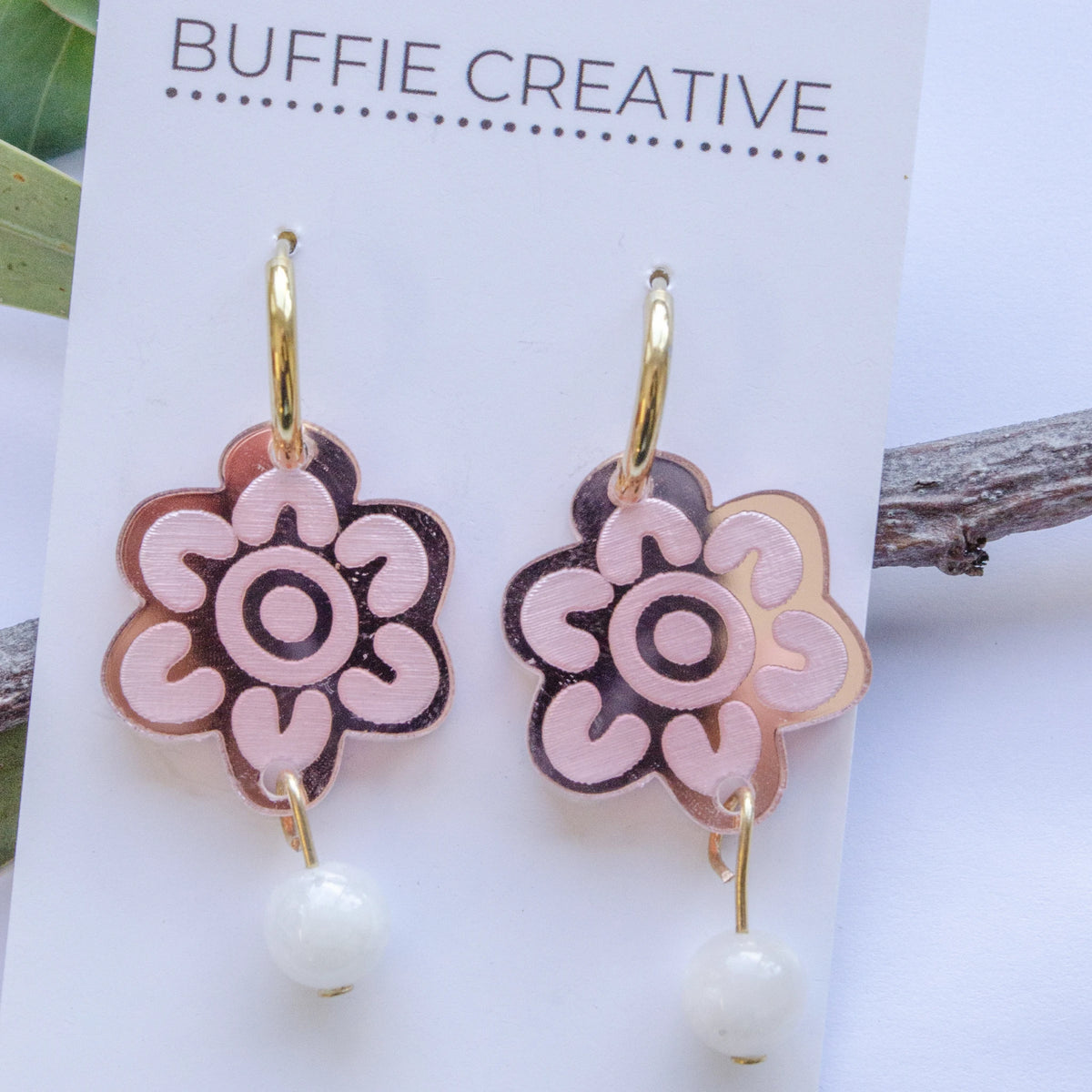 Blush Bloom Pearl Drop Earrings on Buffie Creative card packaging, featuring floral flower-shaped charms in blush pink and rose gold with dangling pearl accents on gold-toned hoops, designed by Buffie Punch.