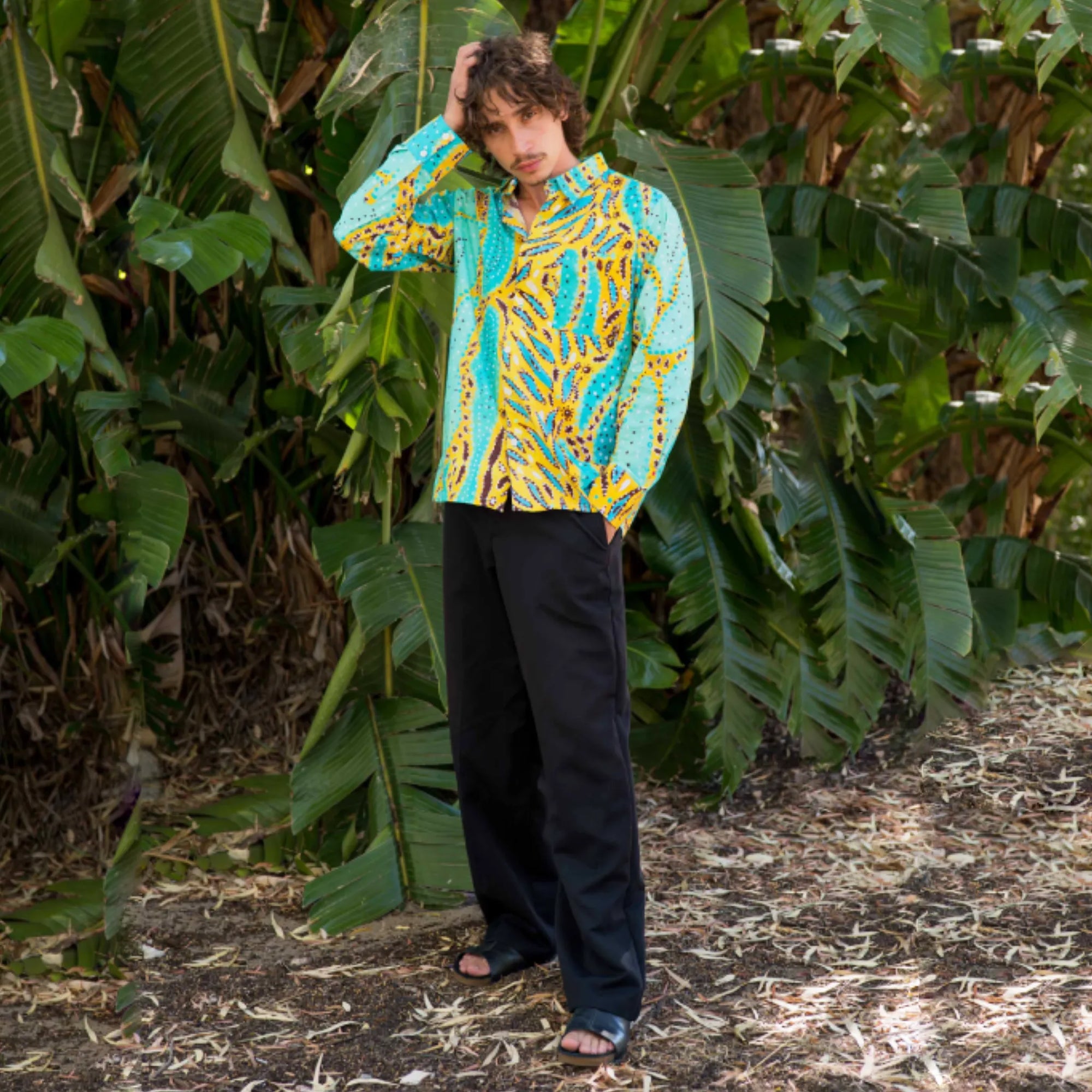 Model wearing the Kirrikin unisex Karijini long-sleeve button-down shirt featuring First Nations artwork against green foliage.