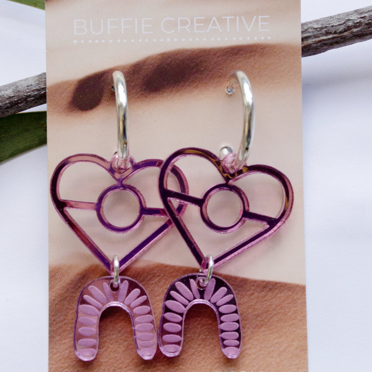 Pink Heart &amp; Arch Statement Earrings on Buffie Creative card packaging, featuring translucent pink heart charms with dangling pattern-etched arch drops on silver-toned hoops, designed by Buffie Punch.